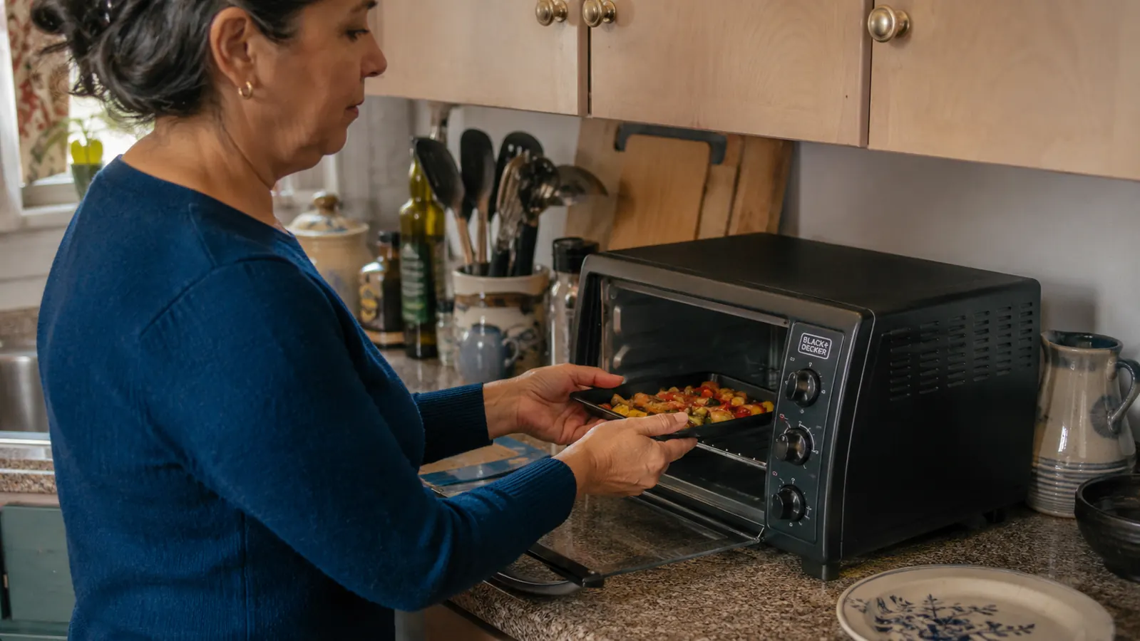 A hand sliding a small baking tray with two chicken thighs into the BLACK+DECKER toaster oven