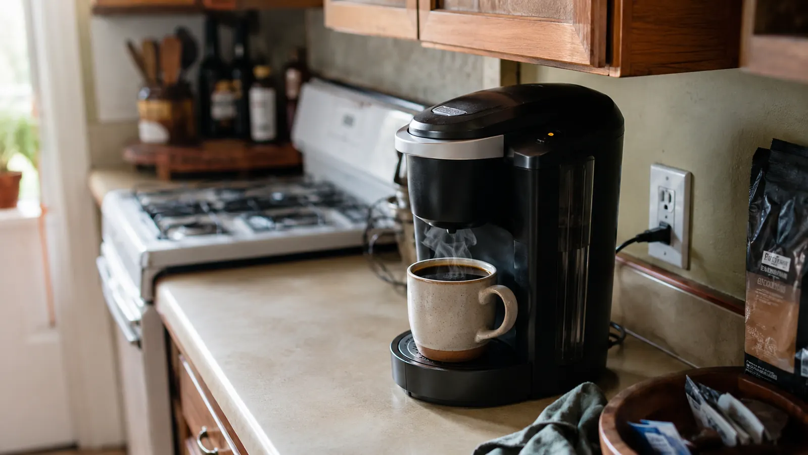 Slim single-serve coffee maker on a narrow kitchen counter with a full dark mug of coffee steaming beside it