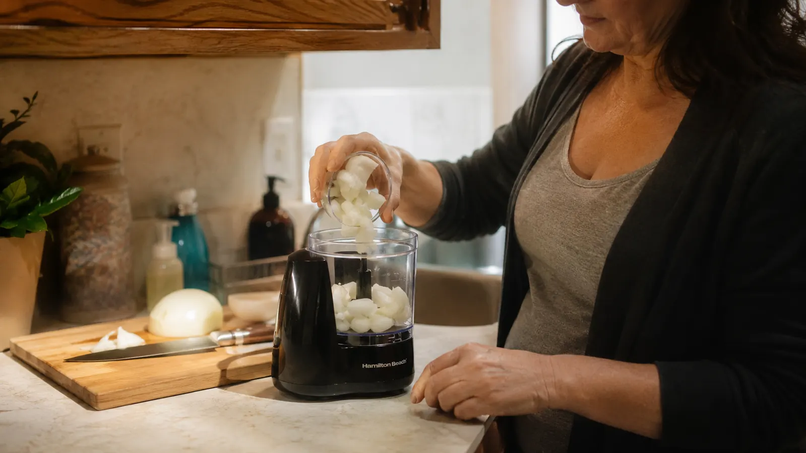Hand dropping quartered onion pieces into the clear plastic bowl of the Hamilton Beach chopper before locking the lid