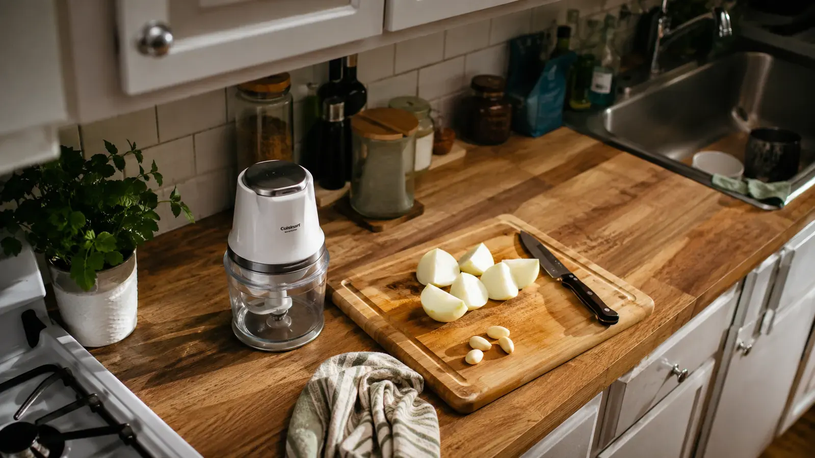Hand loading diced onion and garlic into the clear bowl of a small Hamilton Beach chopper on a narrow kitchen counter