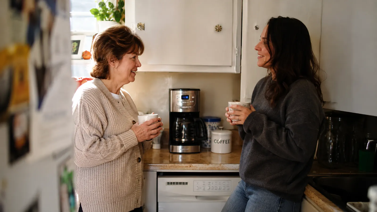 Two women in a small kitchen holding two different mugs of coffee, one decaf and one strong, standing at a corner counter