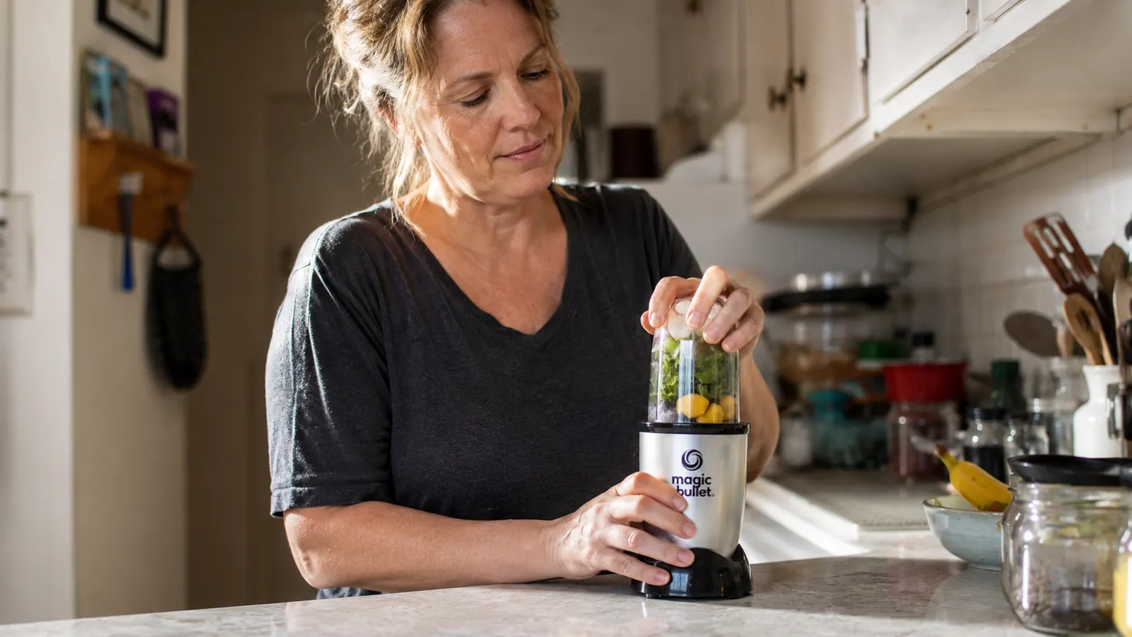 Hands twisting a Magic Bullet blending cup filled with spinach, banana, and frozen mango onto the base